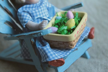 A little porcelain baby doll in blue checkered dress holds an Easter basket with colored eggs for a favorite Christian holiday.