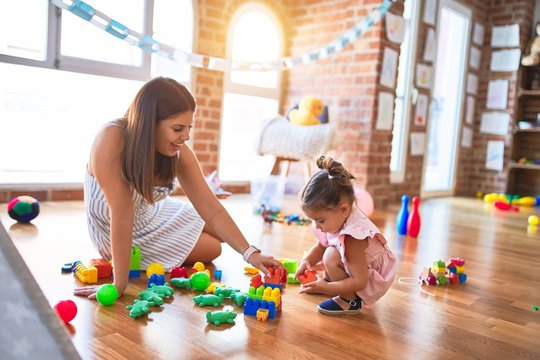 Young beautiful teacher and toddler sitting on the floor playing with building blocks toy at kindergarten
