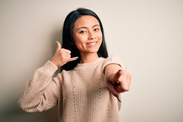 Young beautiful chinese woman wearing casual sweater over isolated white background smiling doing talking on the telephone gesture and pointing to you. Call me.
