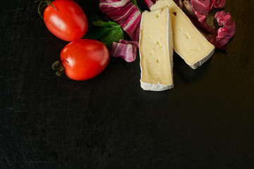Cheese delikatessen closeup on black stone desk surface. Camembert or brie circle in brown kraft paper decorated with basil and pieces of cherry tomatoes, top view image with copy space © Oleg