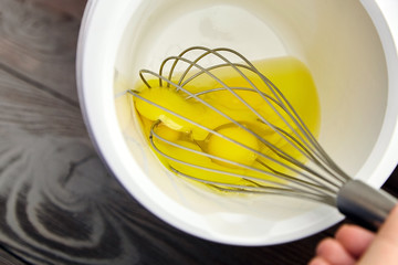 Beating raw yellow egg yolks with a handle steel whisk in white bowl on wooden table. Mixing food...