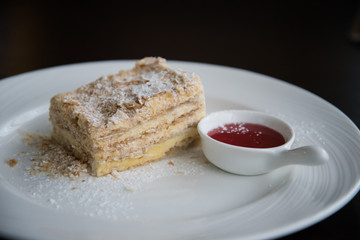 A piece of Napoleon cake on a white plate. The cake is sprinkled with powdered sugar. There is a Cup of jam next to it.
