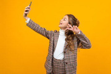 cute schoolgirl in a brown jacket takes a photo with outstretched hand on a smartphone on a yellow background