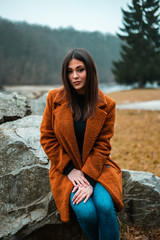 Young attractive female model in brown - orange coat sitting on rocks (stones) in nature. Fashion portrait of beautiful and trendy brunette girl in seasonal clothes (jeans, coat, high heels).