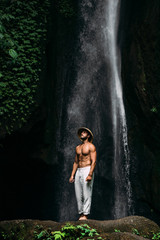 A man of athletic build at the waterfall. A man travels the world. Man at the waterfall. Travel to Bali Indonesia. A man in a conical hat and white pants at the waterfall. Lone traveler.
