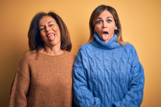 Middle age beautiful couple of sisters wearing casual sweater over isolated yellow background sticking tongue out happy with funny expression. Emotion concept.