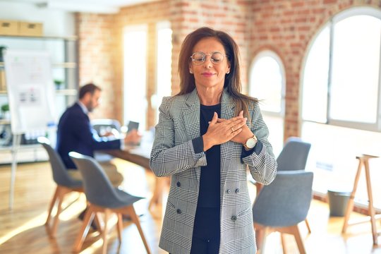 Middle age beautiful businesswoman wearing jacket and glasses standing at the office smiling with hands on chest with closed eyes and grateful gesture on face. Health concept.