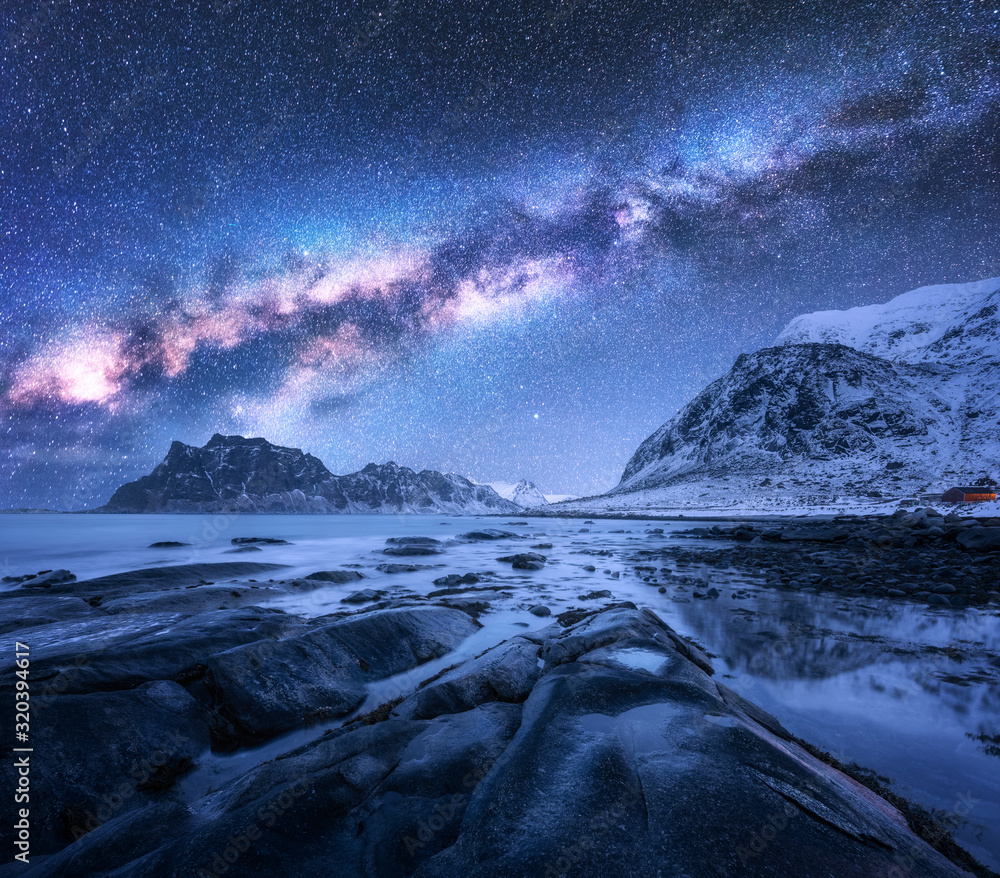 Wall mural milky way above snow covered mountains and rocky beach in winter at night in lofoten islands, norway