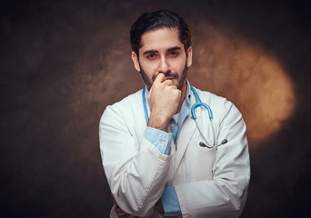 Handsome bearded man in doctor's uniform is posing for photographer over dark background.