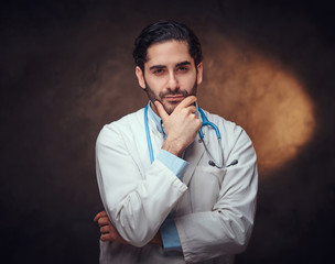 Handsome bearded man in doctor's uniform is posing for photographer over dark background.