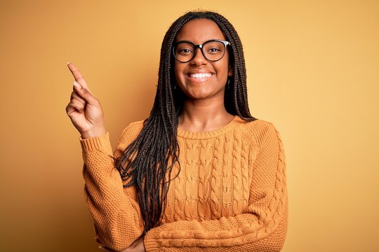 Young african american smart woman wearing glasses and casual sweater over yellow background with a big smile on face, pointing with hand and finger to the side looking at the camera.