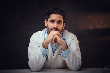 Tired young doctor is sitting next to the table while posing for photographer.