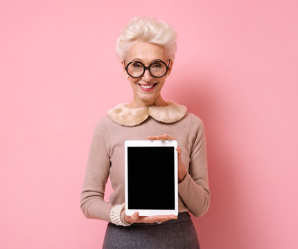 Grandmother holds a digital tablet with a blank screen. Photo of kind elderly woman on pink background.
