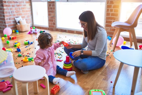 Young beautiful teacher and toddler sitting on the floor building pyramid using hoops at kindergarten