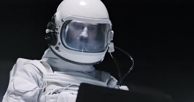 Close up of the male Caucasian astronaut in the helmet from the space costume standing on the Moon at night and using tablet computer.