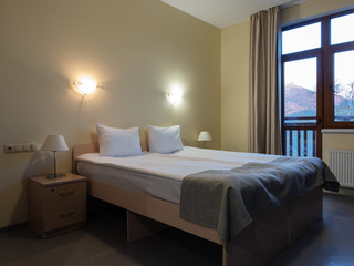 A room with a neatly made double bed, a bedside table with burning lamps and a mountain view from the window