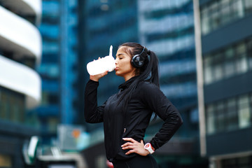 Young woman in black sportswear drinking protein shake after jogging in cold weather holding protein shake and listens to music through headphones