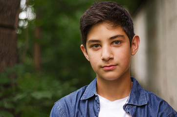 Portrait of a dark-haired teenager in a denim shirt against a background of greenery. The guy looks at the camera. Selective focus. Close up.