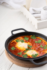 Shakshuka - a dish of eggs fried in a sauce of tomato, hot pepper, onion and seasoning. Shakshuka Scrambled eggs with tomatoes and vegetables on white wooden background. Breakfast in a Frying Pan.