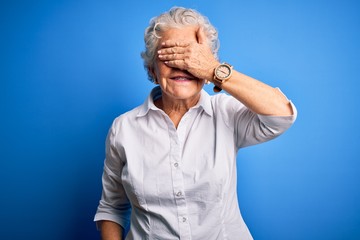 Senior beautiful woman wearing elegant shirt standing over isolated blue background smiling and laughing with hand on face covering eyes for surprise. Blind concept.