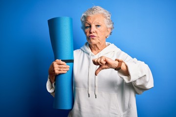 Senior beautiful sporty woman holding mat for yoga standing over isolated blue background with...