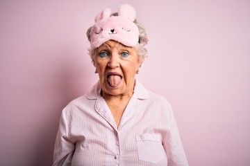 Senior beautiful woman wearing sleep mask and pajama over isolated pink background sticking tongue...