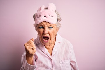 Senior beautiful woman wearing sleep mask and pajama over isolated pink background angry and mad...