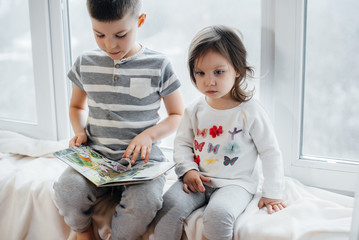 Brother and sister are sitting on the windowsill and reading a book. Happiness, family