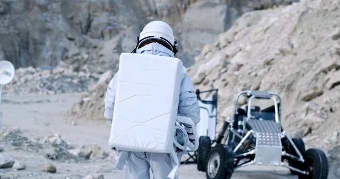 Back view on the NASA spaceman in the full equipped armor walking towards moonwalker in the Moon dessert. Rear. Outside.