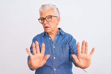 Senior grey-haired woman wearing denim shirt and glasses over isolated white background Moving away hands palms showing refusal and denial with afraid and disgusting expression. Stop and forbidden.