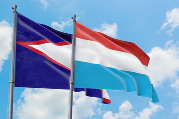 Luxembourg and American Samoa flags waving in the wind against white cloudy blue sky together....