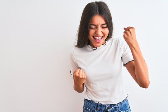 Young beautiful woman wearing casual t-shirt standing over isolated white background very happy and excited doing winner gesture with arms raised, smiling and screaming for success. Celebration