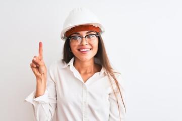 Young beautiful architect woman wearing helmet and glasses over isolated white background showing and pointing up with finger number one while smiling confident and happy.