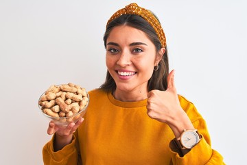 Young beautiful woman holding bowl with peanuts standing over isolated white background happy with big smile doing ok sign, thumb up with fingers, excellent sign