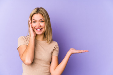 Young blonde caucasian woman isolated holds copy space on a palm, keep hand over cheek. Amazed and...