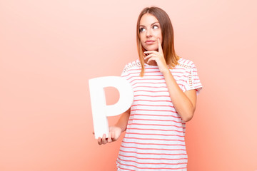 young pretty red head woman confused, doubtful, thinking, holding the letter P of the alphabet to...