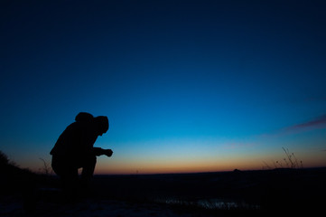 Pray. Repentance. Silhouetted men on a background of blue sky and sunset. Kneeling Prayer to God. Glorification. Praising God - obrazy, fototapety, plakaty