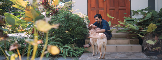 A man with dogs sits on the porch of a house in a tropical garden.