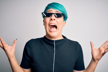 Young woman with blue fashion hair wearing thug life sunglasses over white background crazy and mad shouting and yelling with aggressive expression and arms raised. Frustration concept.