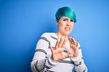 Young beautiful woman with blue fashion hair wearing casual sweater standing at studio disgusted expression, displeased and fearful doing disgust face because aversion reaction. With hands raised