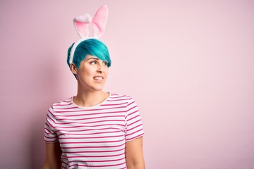 Young woman with fashion blue hair wearing easter rabbit ears over pink background looking away to side with smile on face, natural expression. Laughing confident.