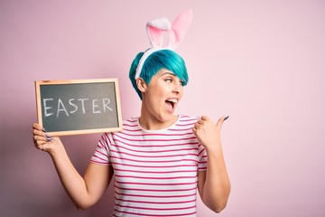 Young woman with fashion blue hair wearing easter rabbit ears and holding blackboard with holiday word pointing and showing with thumb up to the side with happy face smiling