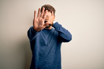 Young handsome man with beard wearing casual sweater standing over white background covering eyes with hands and doing stop gesture with sad and fear expression. Embarrassed and negative concept.