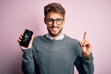 Young man with beard wearing glasses holding broken and craked smartphone surprised with an idea or...