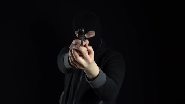 A man in a balaclava mask stands with a gun. The thug points his gun and shoots at the camera. On a black background.