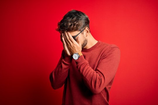 Young handsome man with beard wearing glasses and sweater standing over red background with sad expression covering face with hands while crying. Depression concept.