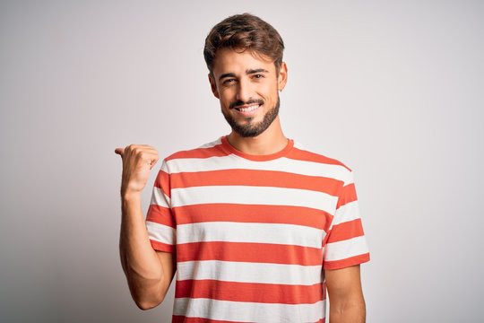 Young handsome man with beard wearing striped t-shirt standing over white background smiling with happy face looking and pointing to the side with thumb up.