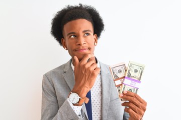 Young african american businessman holding dollars standing over isolated white background serious...