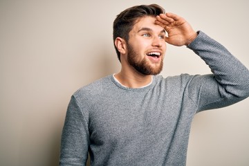 Young handsome blond man with beard and blue eyes wearing casual sweater very happy and smiling looking far away with hand over head. Searching concept.