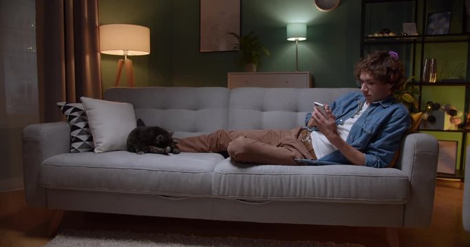 Young Caucasian guy tapping and texting on the smartphone while lying on the couch in dark room with a cat beside. At night. Indoor.
